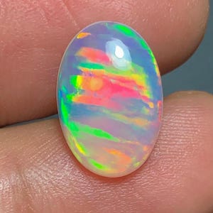May include: An oval-shaped opal gemstone with a vibrant display of colors. The stone showcases a mix of pink, green, yellow, and orange hues, creating a rainbow effect. The opal is set against a neutral background.