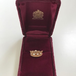Op de afbeelding: Een goudkleurige kroonring met parelaccenten, tentoongesteld in een open bordeauxrode fluwelen sieradendoos. Het ingewikkelde ontwerp van de ring is zichtbaar. De doos heeft een gouden embleem aan de binnenkant.