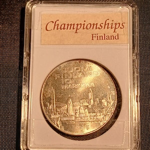 May include: A gold-colored coin from Finland, encased in a clear plastic holder. The coin features a detailed cityscape and the text "SUOMI FINLAND 10 MARKKA". The holder has a label that reads "Championships Finland".
