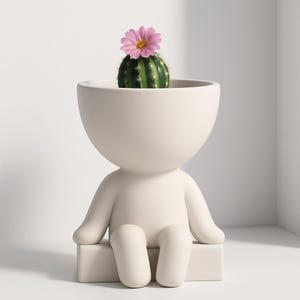 May include: A cream-colored ceramic planter shaped like a person, with arms and legs. A small green cactus with a pink flower sits in the head of the planter. The planter is sitting on a cream-colored rectangular base.