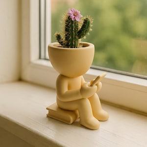 Op de afbeelding: Een gele keramische plantenbak in de vorm van een persoon die een tablet leest. Het hoofd van de figuur is een pot met een groene cactus met een roze bloem. De plantenbak staat op een witte vensterbank.