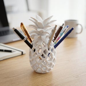 Könnte beinhalten: Ein weißer Stifthalter in Form einer Ananas mit Wabenmuster, der verschiedene Stifte enthält. Der Stifthalter steht auf einem Holztisch, im Hintergrund ein Laptop und ein Notizbuch. Der Stifthalter ist etwa 15 cm hoch.