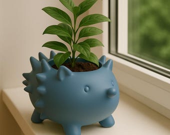 Puede incluir: Una maceta azul con forma de erizo con una pequeña planta verde. La maceta tiene un diseño puntiagudo alrededor del borde y patas pequeñas. La planta tiene varias hojas verdes y está plantada en tierra marrón.