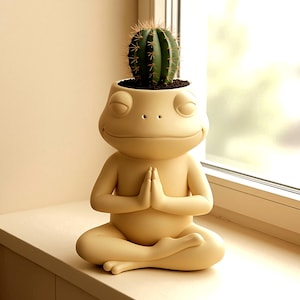 Puede incluir: Una maceta en forma de rana de color amarillo claro, en posición de yoga sentado, con las manos juntas en oración. Un cactus verde con espinas está plantado en la cabeza de la rana. La maceta está sobre una superficie blanca cerca de una ventana.
