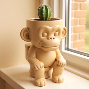 Op de afbeelding: Een bruin/beige aapvormige plantenbak met een groene cactus. De plantenbak heeft een gedetailleerd apengezicht, oren en armen. De cactus is geplant in donkerbruine aarde. De plantenbak staat op een witte ondergrond.