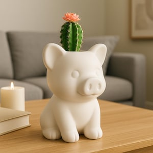 May include: A white ceramic pig-shaped planter holding a green cactus with a pink flower. The planter has a smiling face and is sitting on a wooden table. A lit candle and a book are in the background.