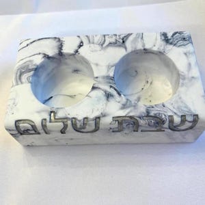 May include: A rectangular white candle holder with a black marbled pattern featuring two circular indentations for candles. The front side displays the Hebrew text Shabbat Shalom engraved in a dark, metallic-toned finish across the center.