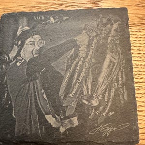 May include: A square slate coaster featuring a laser-engraved image of a person holding a trophy. The design includes the text Premier League repeated multiple times and a signature at the bottom right corner. The coaster has a rough, natural edge.