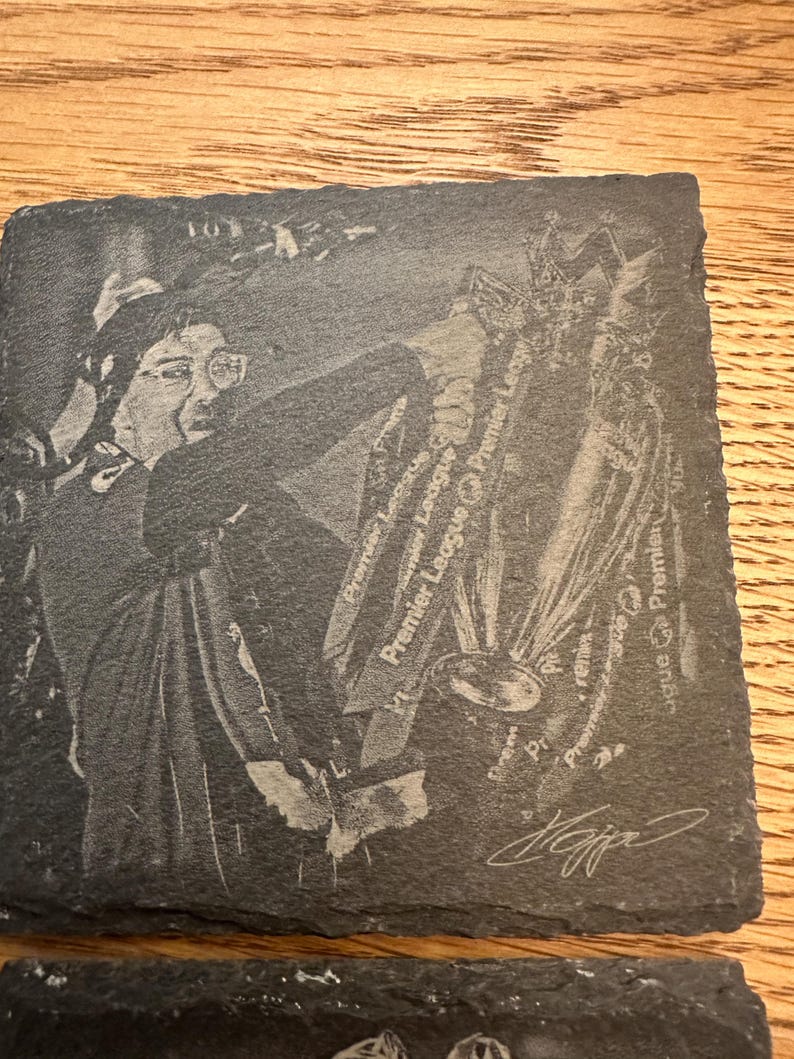 May include: A square slate coaster featuring a laser-engraved image of a person holding a trophy. The design includes the text Premier League repeated multiple times and a signature at the bottom right corner. The coaster has a rough, natural edge.