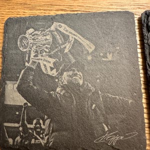 May include: A square dark gray slate coaster featuring a laser-engraved image of a person holding a trophy overhead. The trophy has the text Energy Drink and Carabao visible. A signature is engraved in the bottom right corner of the coaster.