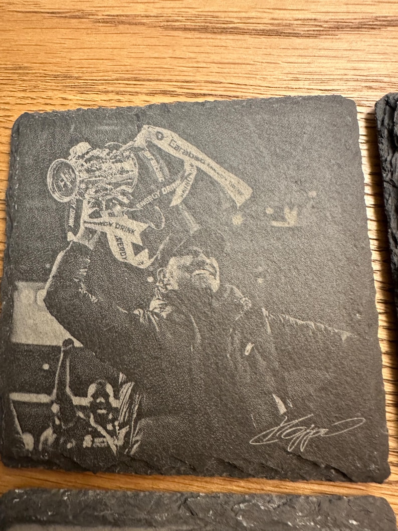 May include: A square dark gray slate coaster featuring a laser-engraved image of a person holding a trophy overhead. The trophy has the text Energy Drink and Carabao visible. A signature is engraved in the bottom right corner of the coaster.