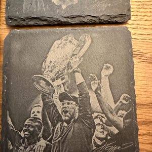 May include: A square slate coaster featuring a laser-engraved image of a person holding a large trophy overhead, surrounded by other individuals with their arms raised in celebration. The design includes a signature in the bottom right corner.