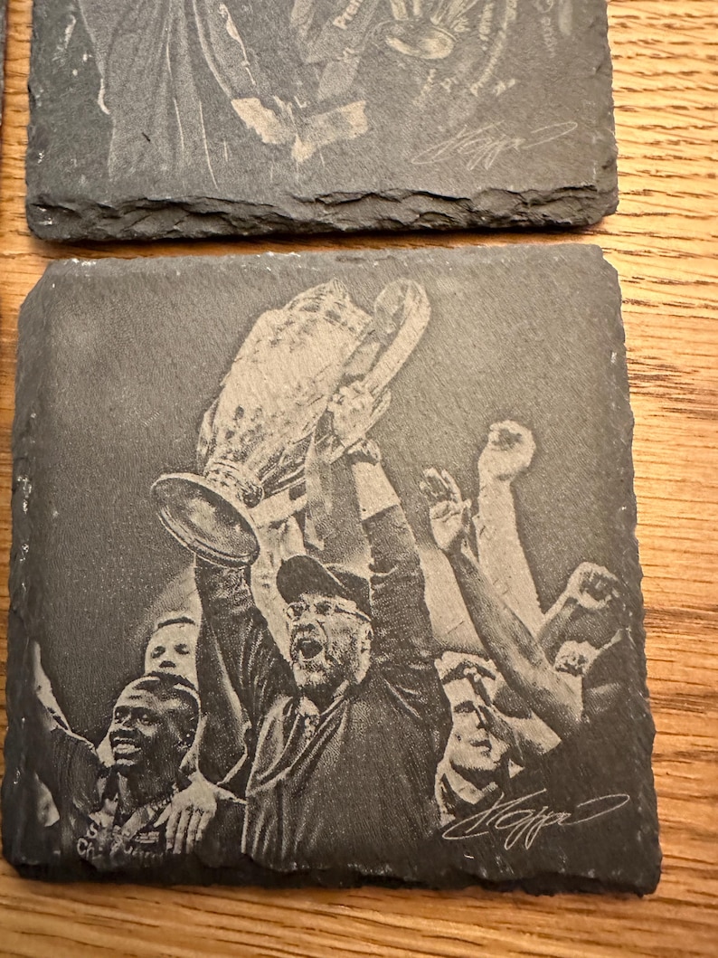 May include: A square slate coaster featuring a laser-engraved image of a person holding a large trophy overhead, surrounded by other individuals with their arms raised in celebration. The design includes a signature in the bottom right corner.