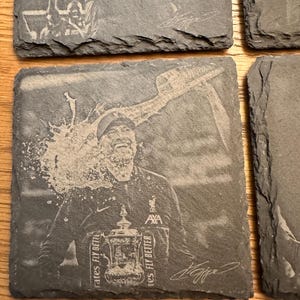 May include: A square dark gray slate coaster featuring a laser-engraved image of a smiling person wearing a jacket with the AXA logo, holding a trophy with the text Emirates Fly Better visible, with a liquid splash effect and a signature.