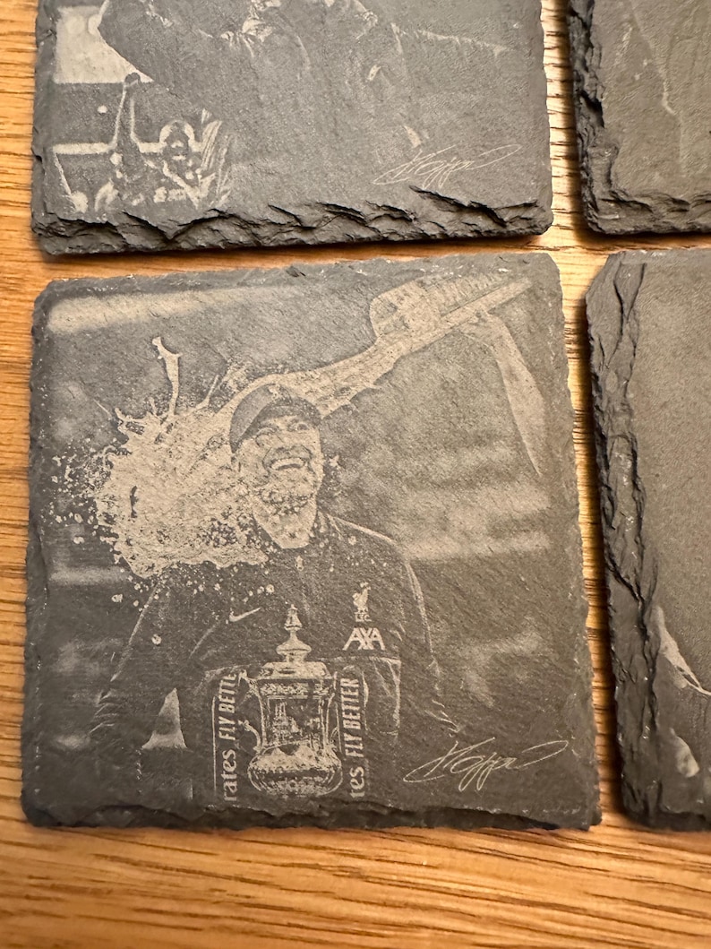 May include: A square dark gray slate coaster featuring a laser-engraved image of a smiling person wearing a jacket with the AXA logo, holding a trophy with the text Emirates Fly Better visible, with a liquid splash effect and a signature.
