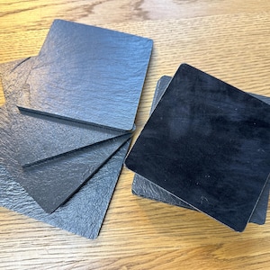 May include: A set of square coasters featuring a dark gray, textured slate surface on one side and a smooth, solid black finish on the reverse. The coasters are arranged in a small stack and a fanned-out group, showcasing their dual-sided design.