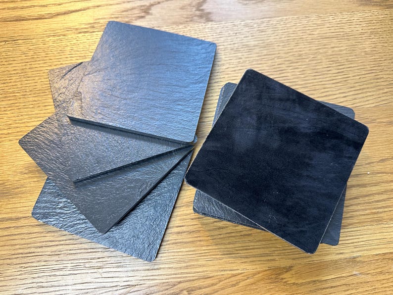 May include: A set of square coasters featuring a dark gray, textured slate surface on one side and a smooth, solid black finish on the reverse. The coasters are arranged in a small stack and a fanned-out group, showcasing their dual-sided design.