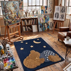 May include: A navy blue rug featuring a sleeping teddy bear design with a crescent moon and star accents. The rug is in a room with art supplies, paintings, and easels, suggesting an art studio setting.