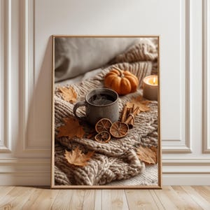 Puede incluir: Un cuadro decorativo enmarcado que muestra una taza de cerámica gris con una bebida oscura humeante, una pequeña calabaza naranja, rodajas de naranja seca, ramas de canela y hojas de otoño marrones sobre una manta de punto color beige.
