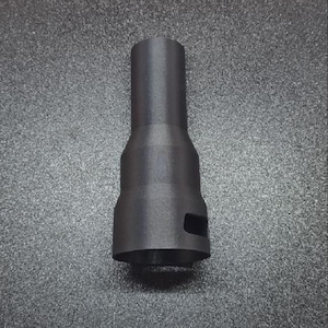 Könnte beinhalten: Ein schwarzer, zylindrischer Adapter mit verbreiterter Basis und schmalerem Oberteil. Der Adapter hat einen rechteckigen Ausschnitt nahe der Basis. Das Objekt besteht aus mattem Material und liegt auf einer strukturierten Oberfläche.