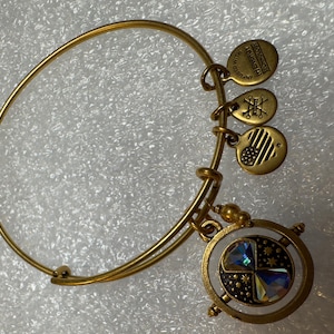 Può includere: Bracciale rigido dorato con tre piccoli ciondoli e un ciondolo più grande e rotondo con un centro di cristallo. Il bracciale ha un design semplice ed elegante, adatto all'uso quotidiano.