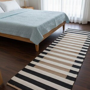 May include: A striped rug in black, beige, and white, placed on a dark wooden floor. The rug is in front of a bed with a light blue quilt. The room has a minimalist design. The rug's pattern is made of horizontal stripes.