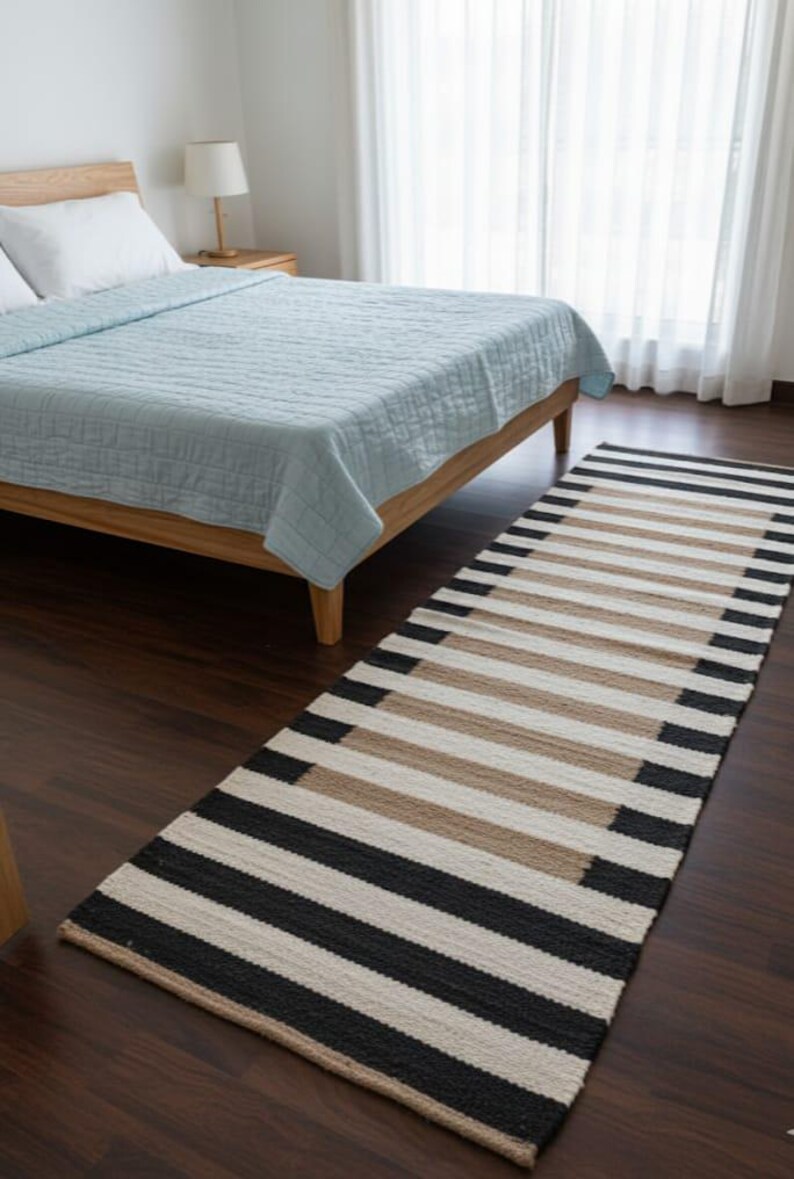May include: A striped rug in black, beige, and white, placed on a dark wooden floor. The rug is in front of a bed with a light blue quilt. The room has a minimalist design. The rug's pattern is made of horizontal stripes.