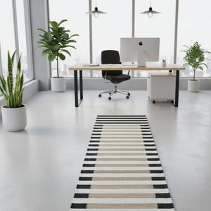 May include: An office interior with a striped rug in black, beige, and white. The room includes a wooden desk, black chair, and potted plants. Large windows provide natural light and a view.