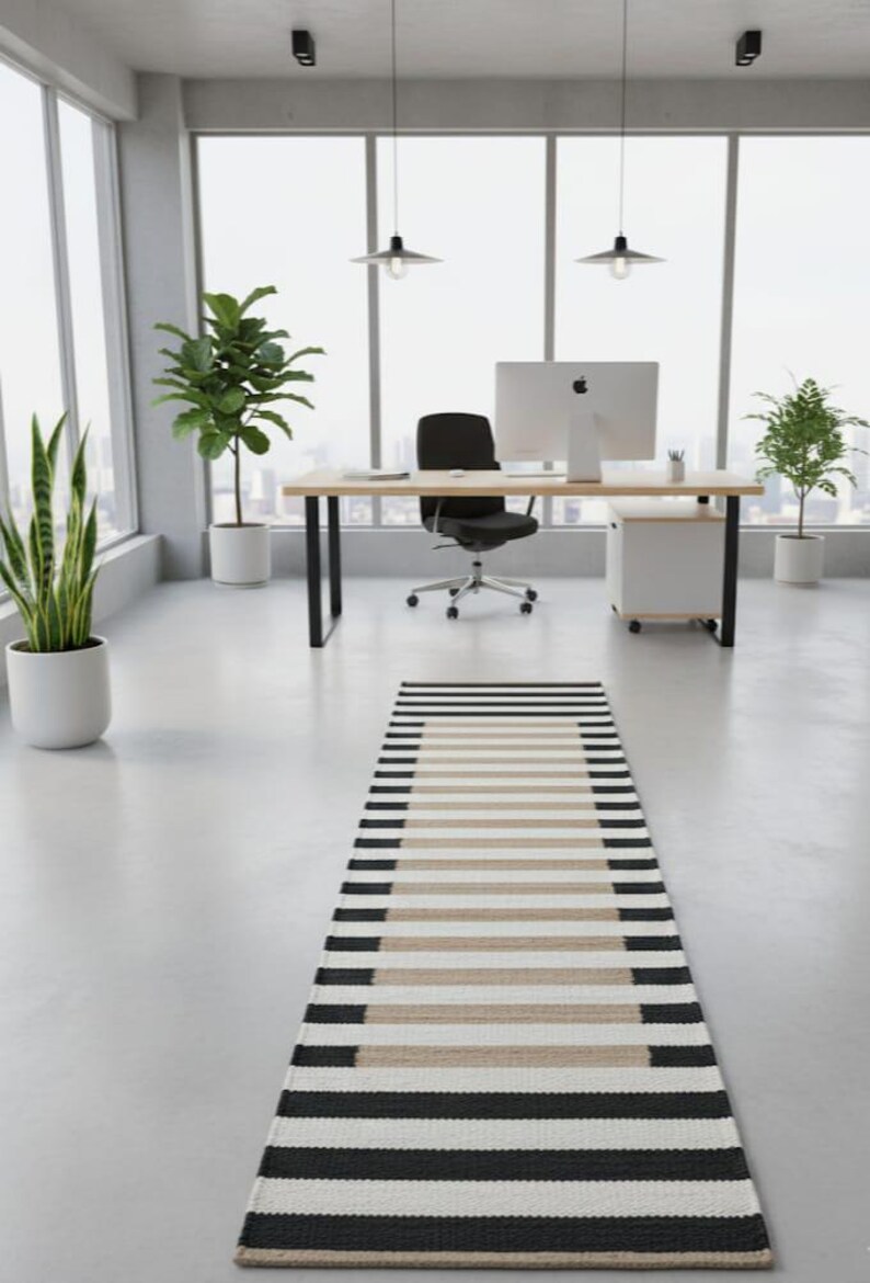 May include: An office interior with a striped rug in black, beige, and white. The room includes a wooden desk, black chair, and potted plants. Large windows provide natural light and a view.