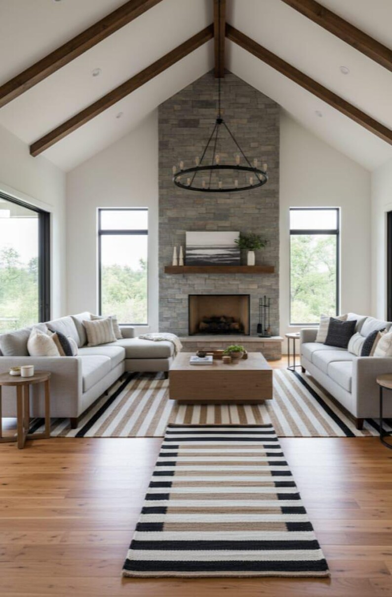 May include: A contemporary living room with a stone fireplace, two light grey sofas, and a wooden coffee table. A striped rug and hardwood floors add warmth. The room features large windows and a chandelier.