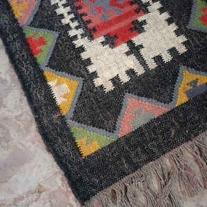 Puede incluir: Una alfombra rectangular tejida a mano con fondo negro y patrones geométricos en rojo, blanco, amarillo, verde y gris. La alfombra tiene un borde con flecos y una apariencia texturizada, adecuada para la decoración del hogar.