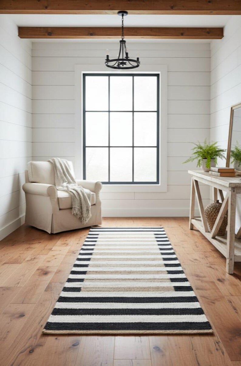 May include: A room with a striped rug in black, beige, and white. There is a cream armchair, a wooden console table, and a large window with a black frame. A chandelier hangs from the ceiling. The room has a neutral colour scheme.