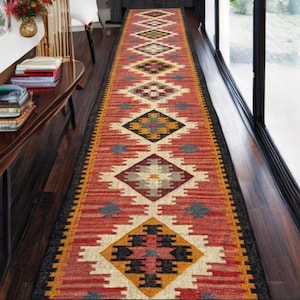 Può includere: Un lungo tappeto runner rettangolare con un motivo geometrico. Il tappeto presenta un motivo centrale di forme a diamante ripetute in rosso, giallo, nero e grigio, bordato da un bordo nero. Il tappeto è posto su un pavimento in legno scuro.