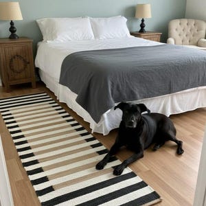 May include: A bedroom scene with a black dog lying on a wooden floor next to a striped rug. The rug has black, beige, and white stripes. A bed with white bedding and a grey blanket is in the background.