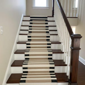May include: A staircase with dark wood steps and white risers. A runner rug with horizontal stripes in black, tan, and cream colours runs up the centre of the stairs. A white banister with a dark wood handrail is on the right.