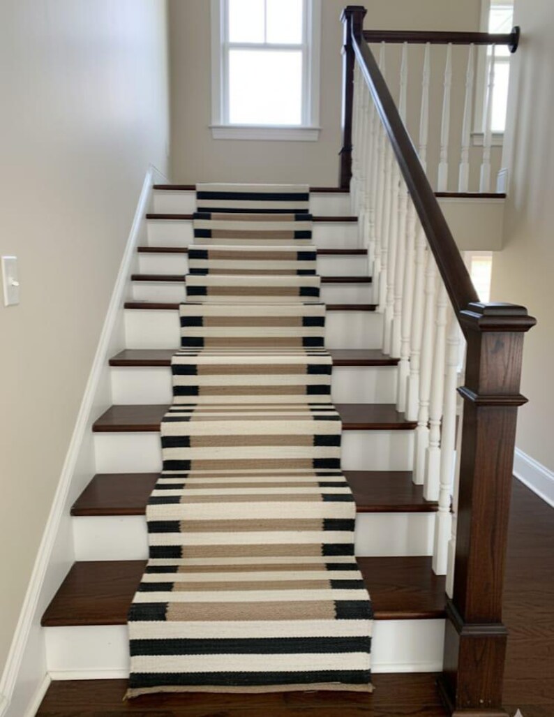 May include: A staircase with dark wood steps and white risers. A runner rug with horizontal stripes in black, tan, and cream colours runs up the centre of the stairs. A white banister with a dark wood handrail is on the right.