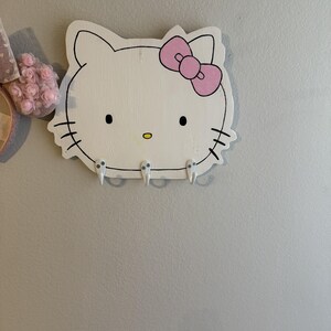 Puede incluir: Un colgador de pared de madera blanca con forma de cabeza de Hello Kitty, con un lazo rosa, ojos negros y bigotes. Tiene tres pequeños ganchos debajo. El diseño está delineado en negro, con una nariz amarilla.
