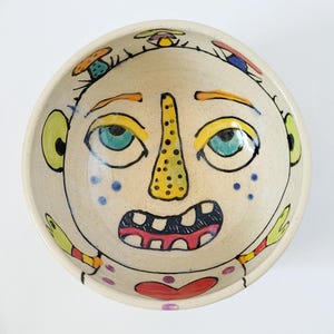 May include: A white ceramic bowl with a hand-painted design of a face with a yellow nose, blue eyes, and a red mouth. The face is surrounded by colorful mushrooms and dots.