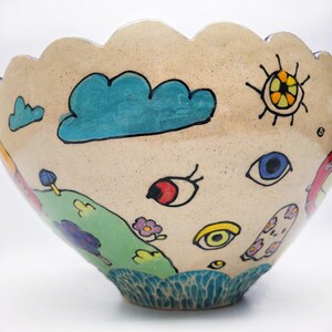 May include: A large ceramic bowl with a scalloped rim and a whimsical design. The bowl is decorated with hand-painted clouds, eyes, and a sun. The bowl is a light beige color with accents of blue, green, yellow, red, and orange.