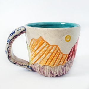 May include: A ceramic mug with a hand-painted design of a mountain range with a yellow sun. The mug has a teal blue interior and a colorful, textured handle.