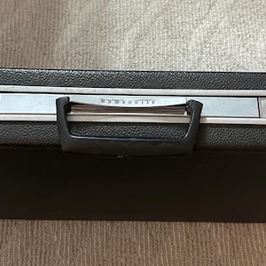 May include: A black Samsonite briefcase with a silver-colored trim and handle. The briefcase has a textured surface and a lock on each side. The handle is black and the brand name is visible on the front.