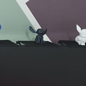 Könnte beinhalten: Drei kleine Figuren: ein blauer Elefant, eine dunkelgraue, außerirdisch aussehende Kreatur und ein weißes Kaninchen. Die Figuren stehen auf einer schwarzen Oberfläche vor einem Hintergrund aus geometrischen Formen in Grau-, Lila- und Weißtönen. Der Elefant ist blau.