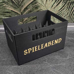 Könnte beinhalten: Eine rechteckige schwarze Aufbewahrungsbox mit Griffaussparungen an jeder Seite. Auf der Vorderseite befindet sich der Schriftzug SPIELEABEND in goldener Farbe. Das Innere verfügt über ein Gitter aus vertikalen Trennwänden für Fächer.