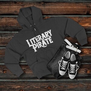 以下が含まれることがあります： 白い「LITERARY PIRATE」の文字が入った濃いグレーのジップアップパーカー、黒のジーンズ、白いベルト、白と黒のスニーカー。木製の表面に配置されています。