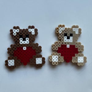 Puede incluir: Dos figuras de osos de peluche hechas a mano con cuentas de plástico de colores. Uno es marrón con un corazón rojo, el otro es de color canela con un corazón rojo. Ambos tienen detalles blancos y están sentados.