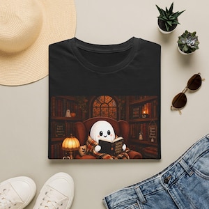 Peut inclure: Un t-shirt noir avec une illustration représentant un fantôme blanc assis dans un fauteuil marron, lisant un livre intitulé Good Books Spooky Vibes. Le décor montre une bibliothèque avec des étagères, une lampe et une fenêtre.