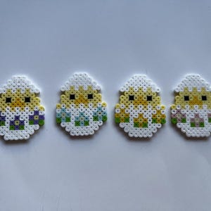 Może przedstawiać: Cztery ręcznie robione dekoracje wielkanocne. Każda z nich przedstawia pisklę wykluwające się z jajka, wykonane z kolorowych plastikowych koralików. Pisklęta są żółte z czarnymi oczami, a jajka białe z kolorowymi akcentami.