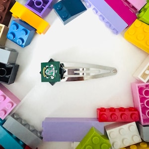May include: A silver metal hair clip with a green and white emblem, surrounded by colorful toy building blocks. The clip features a rectangular emblem with a green background and white detailing. The blocks are various colors, including blue, yellow, and pink.