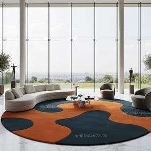 May include: A modern living room featuring a large, round rug with an abstract design in navy blue and orange. The room includes a curved sofa, two armchairs, and a glass coffee table. Two olive trees in pots flank the seating area.