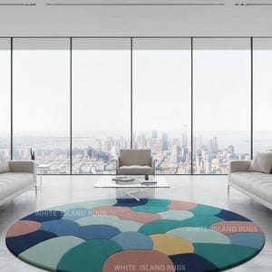 May include: A round area rug with a colorful, abstract design. The rug features a pattern of overlapping, semi-circular shapes in shades of blue, green, pink, and yellow. The words "WHITE ISLAND RUGS" are printed on the rug.